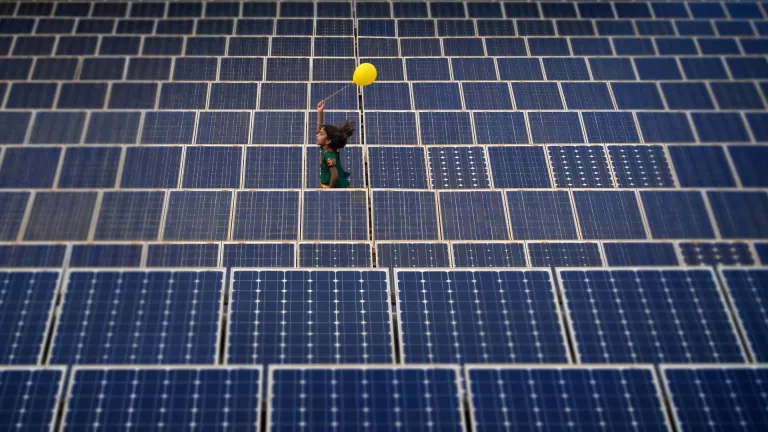 A young girl runs happily amidst rows of solar panels in the background, holding a yellow balloon